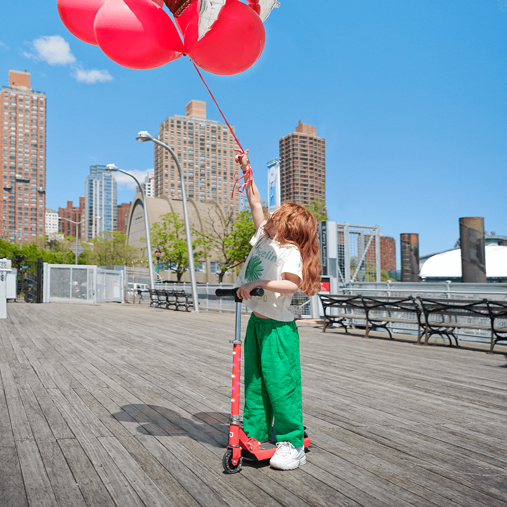 young kid holding onto balloons and a red jupiter kick scooter